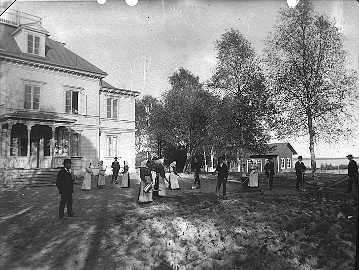Människor i gammeldags kläder framför ett stort hus som påtar i jorden med krattor