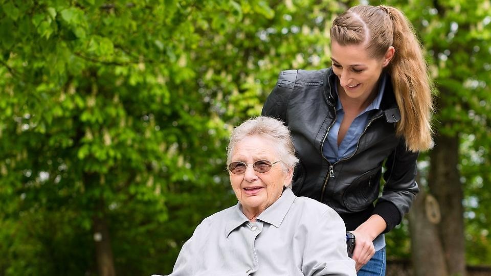 En kvinna leder en äldre dam som sitter i en rullstol