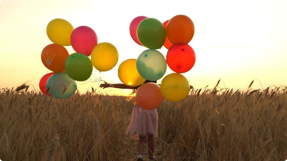 Flicka med färgglada ballonger springer på solig äng.