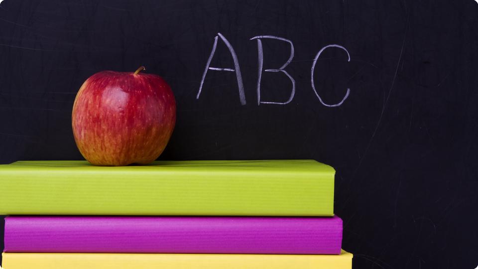 Trave med tre färglada böcker med ett rött äpple på toppen. I bakgrunden syns en griffeltavla med texten ABC. 