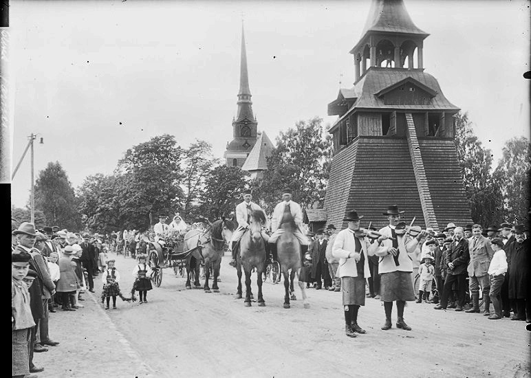Gammal svartvit bild på en parad med hästar och spelmän, framför kyrkan och klockstapeln.