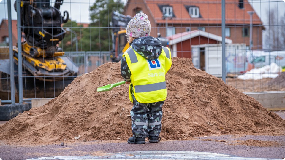 Ett förskolebarn i gul reflexväst som står på en skolgård och håller en grön plastspade i handen, framför en sandhög.